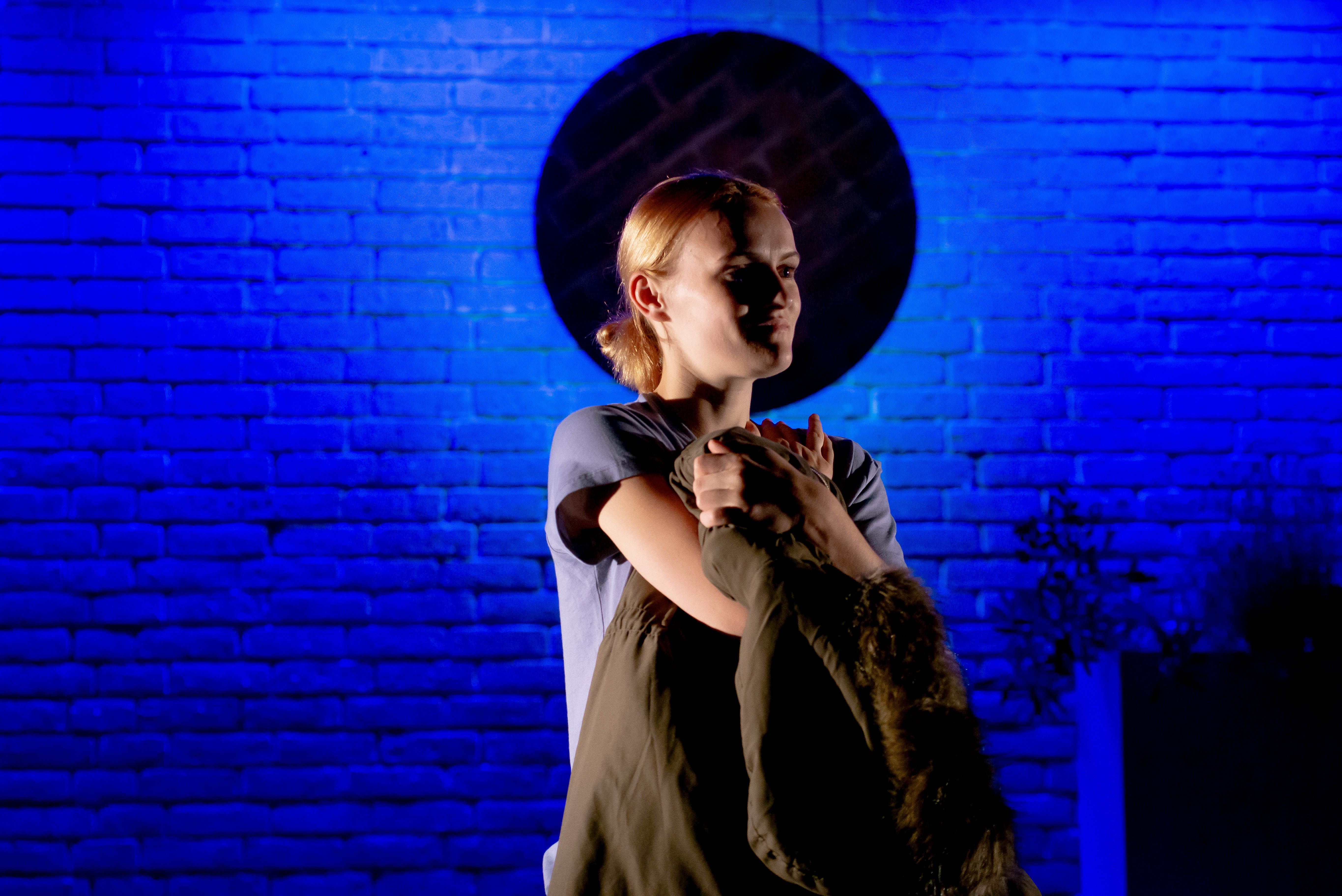 A woman sits on a stage with blue lighting holding a coat or blanket close to their chest. The woman appears thoughtful or somber, illuminated from one side, casting shadows on their face.Photo credit picturesbybish.com